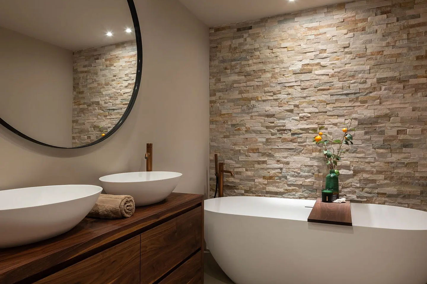 Badkamer met vrijstaand bad, natuurstenen wand en een wastafelmeubel van hout met twee waskommen